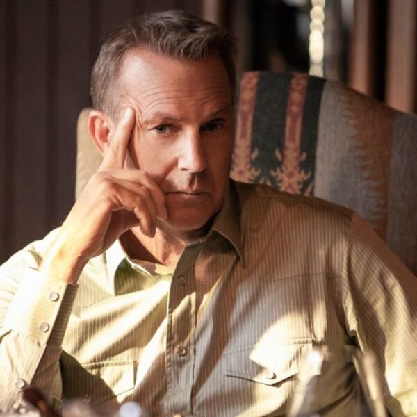 Kevin Costner leaning against his hand while sitting at the dinner table in Yellowstone.